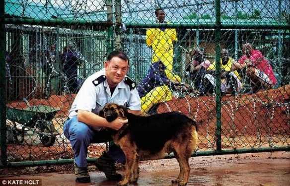 Will Thurbin, former governor of an Isle of Wight prison, poses at Montagne Posse Prison in Seychelles with his dog Lucy, while Somali prisoners behind razor wire look on. Photo by Kate Holt for the Daily Mail